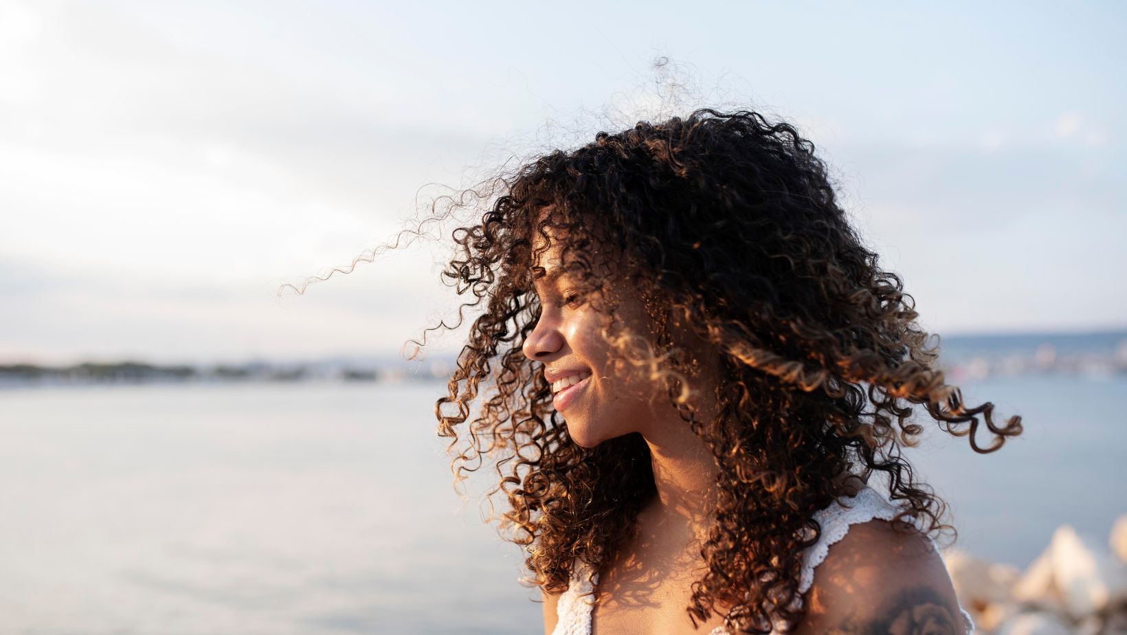 Mulher de cabelo cacheado na praia, exposta ao sol e à água do mar que ressecam os fios