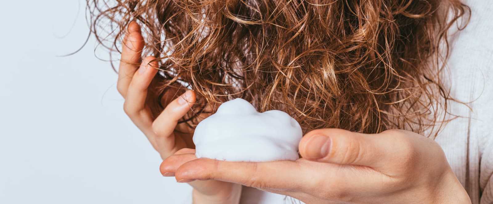 Mão segurando espuma branca de produto capilar diante de cabelo cacheado castanho